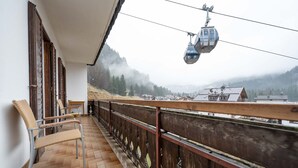 Un balcon avec des fauteuils en osier, une balustrade en bois et une vue sur une ville de montagne, avec un téléphérique visible au loin.