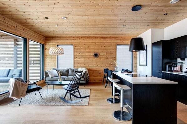 A modern kitchen with a black countertop and bar stools, a wooden ceiling, and a seating area with a sofa and chairs.