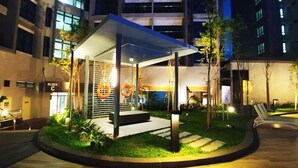 A covered outdoor area with a seating arrangement, surrounded by greenery and a building in the background.