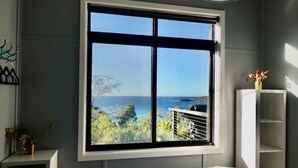 A bedroom with a large window overlooking a coastal view, a bed, a bedside table, and a white shelving unit. - Crow`s Nest - Couples Retreat (Eaglehawk Neck)