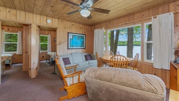 Ein gemütliches Blockhaus-Interieur mit Holzbalkendecke, Deckenventilator, Esstisch mit Stühlen, einer Couch und einem Fenster mit Vorhängen.