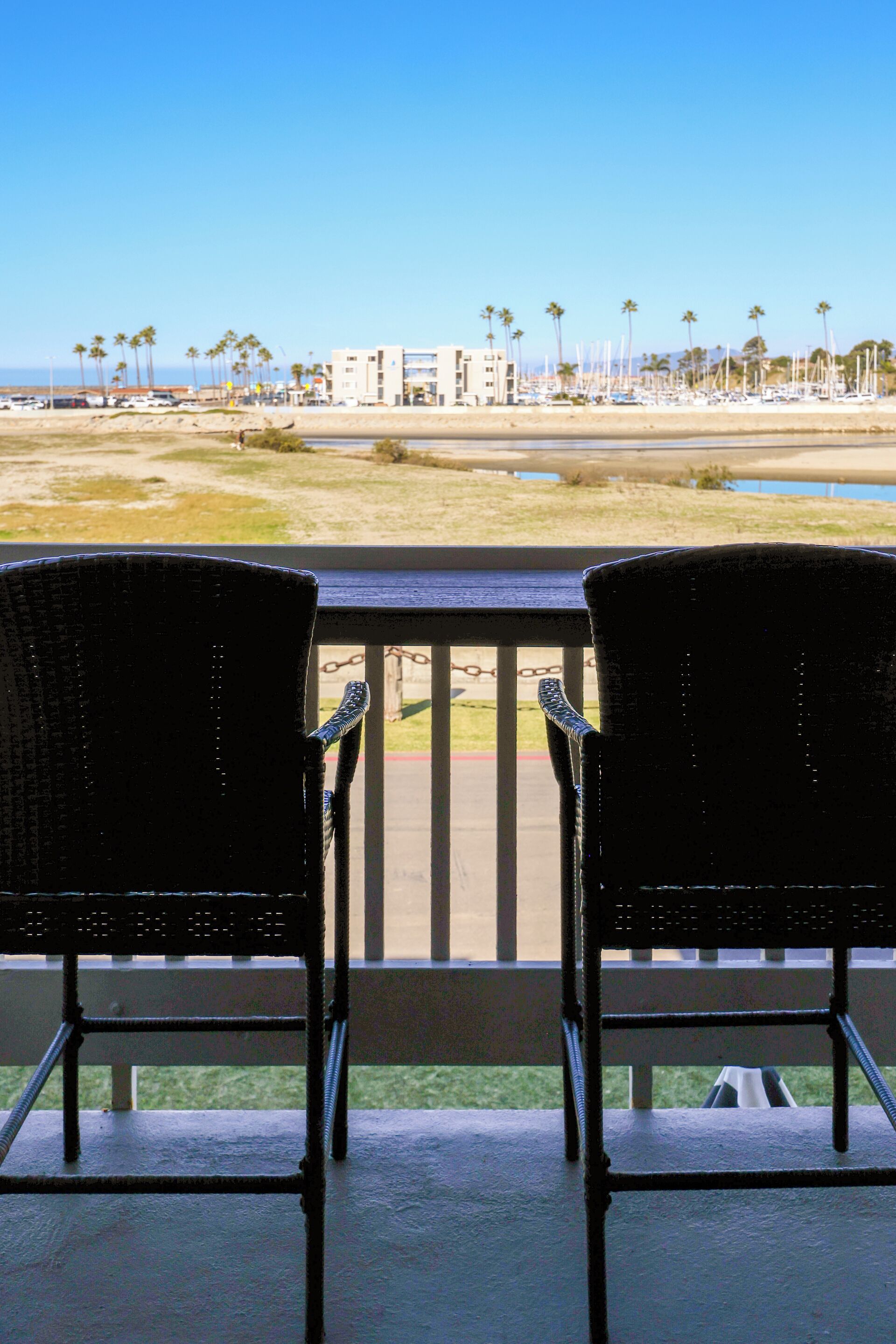 Deux fauteuils en osier noir sur un balcon donnant sur une plage et un port de plaisance.