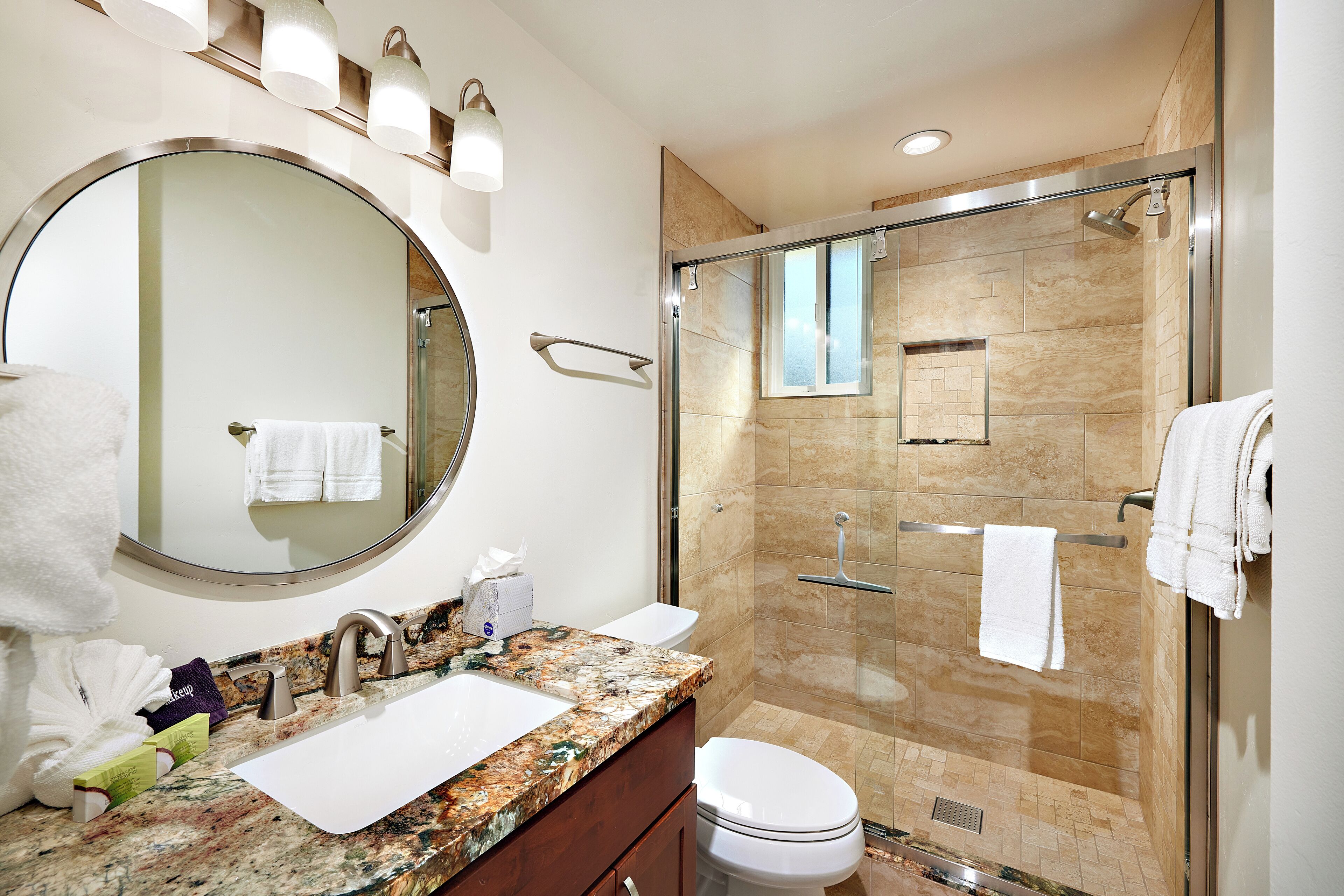 A modern bathroom with a granite countertop, a round mirror, a toilet, and a glass-enclosed shower area.
