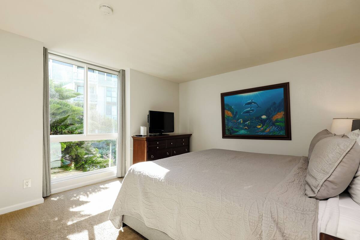 A bedroom with a large window, a bed, a dresser, a television, and an underwater painting on the wall.