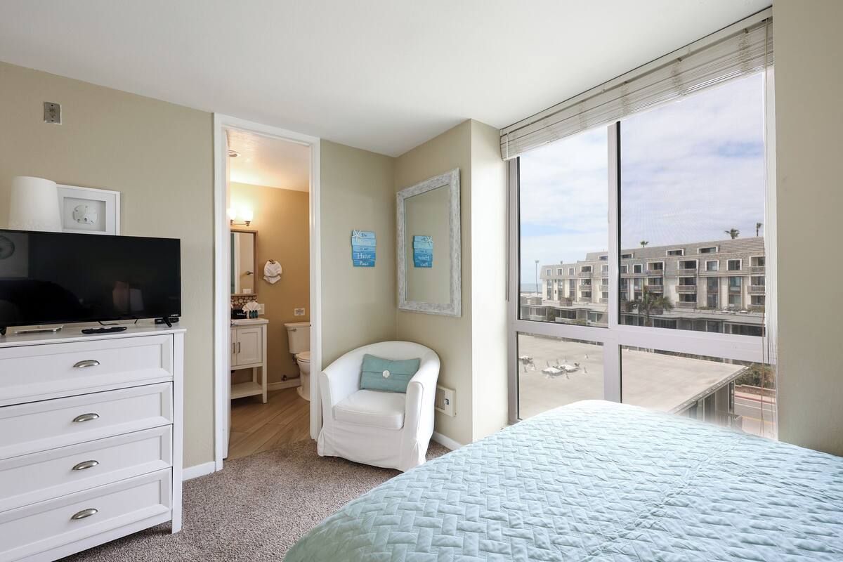 A bedroom with a bed, a TV on a dresser, a chair, a window with a view of buildings, and a bathroom visible through an open door.
