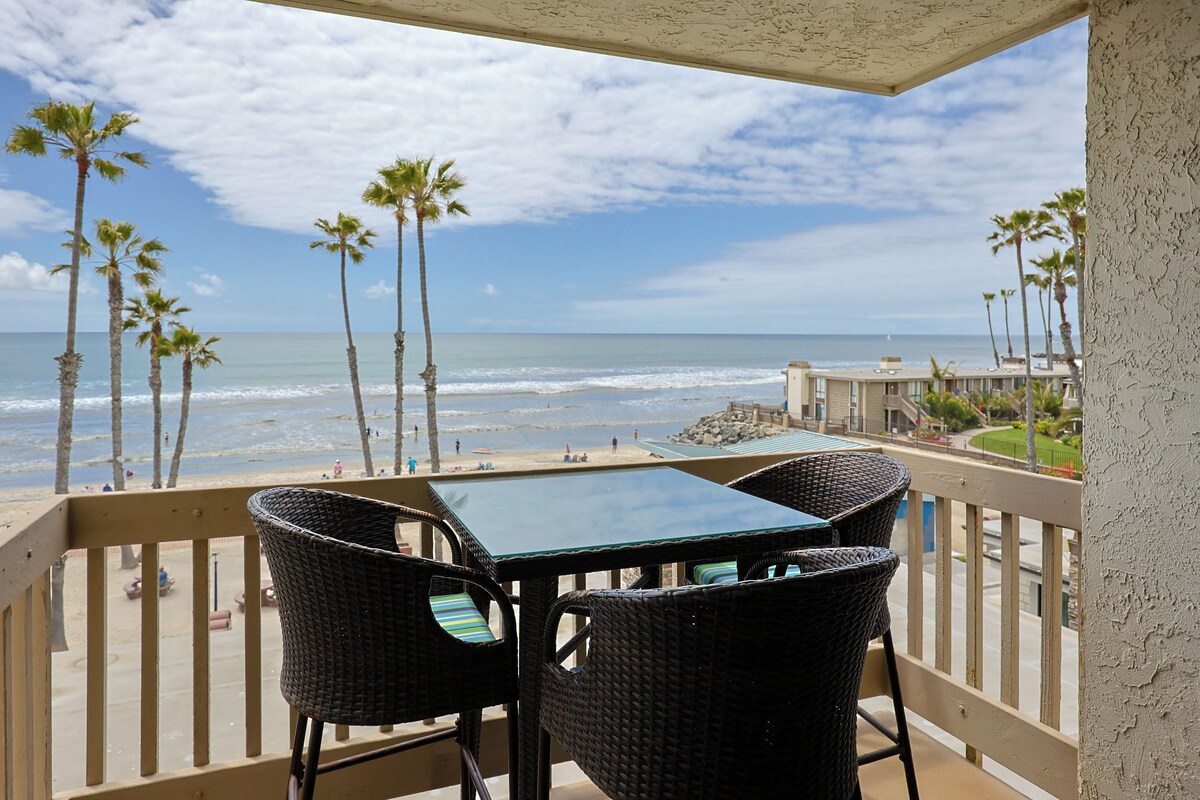 A balcony with wicker chairs and a table overlooking a beach with palm trees and ocean waves.