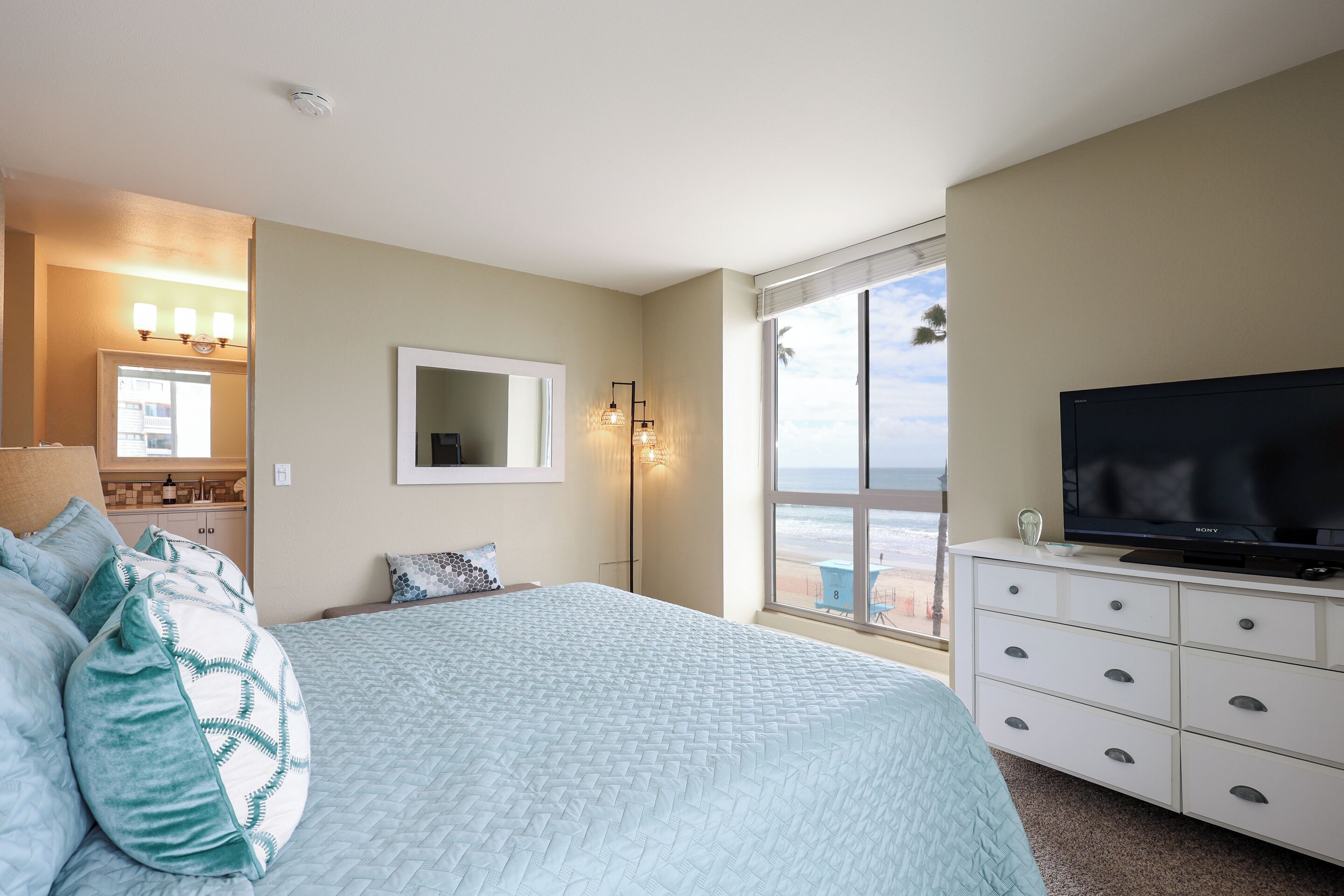 Une chambre à coucher avec un lit, une commode, un téléviseur et une grande fenêtre donnant sur la plage.