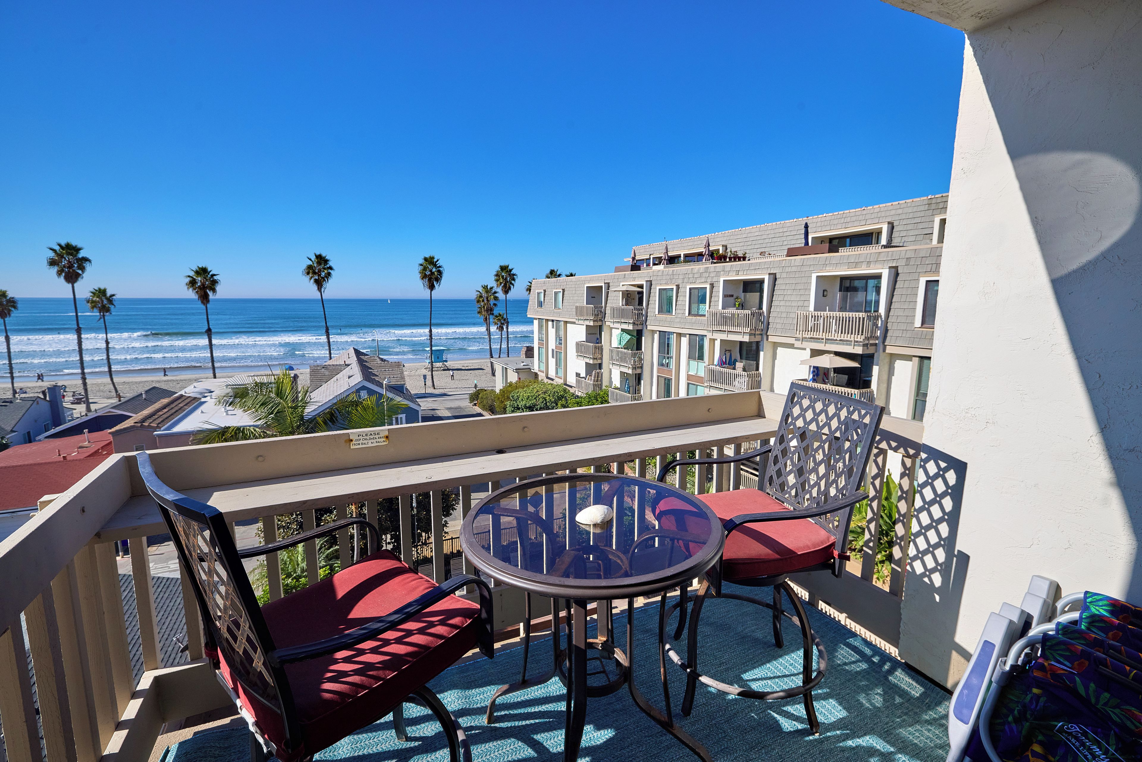 A balcony with a glass table and red cushioned chairs, overlooking a beach and ocean.
