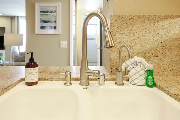 Une salle de bain avec un lavabo, deux robinets, un flacon de gel hydroalcoolique et une bouteille verte de liquide vaisselle.
