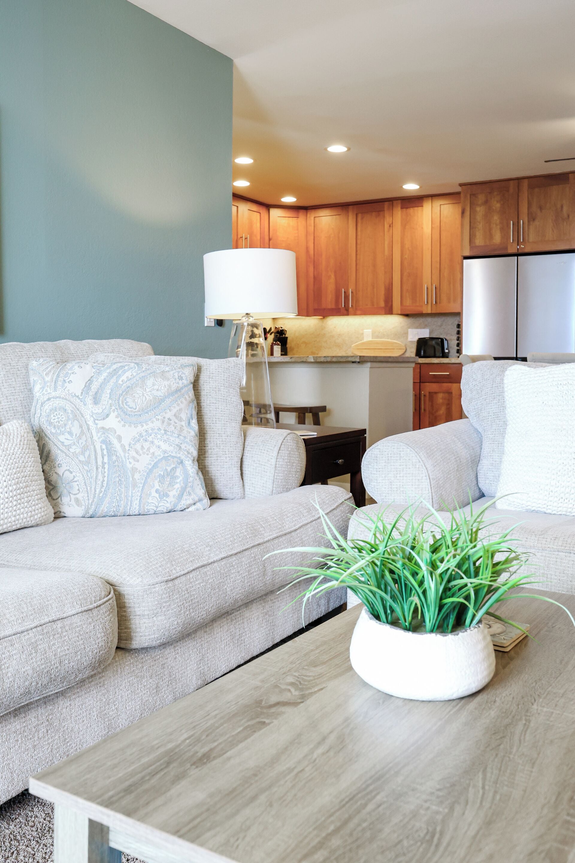 Una sala de estar moderna con un sofá color beige, una mesa de centro blanca con una planta, y una cocina al fondo.