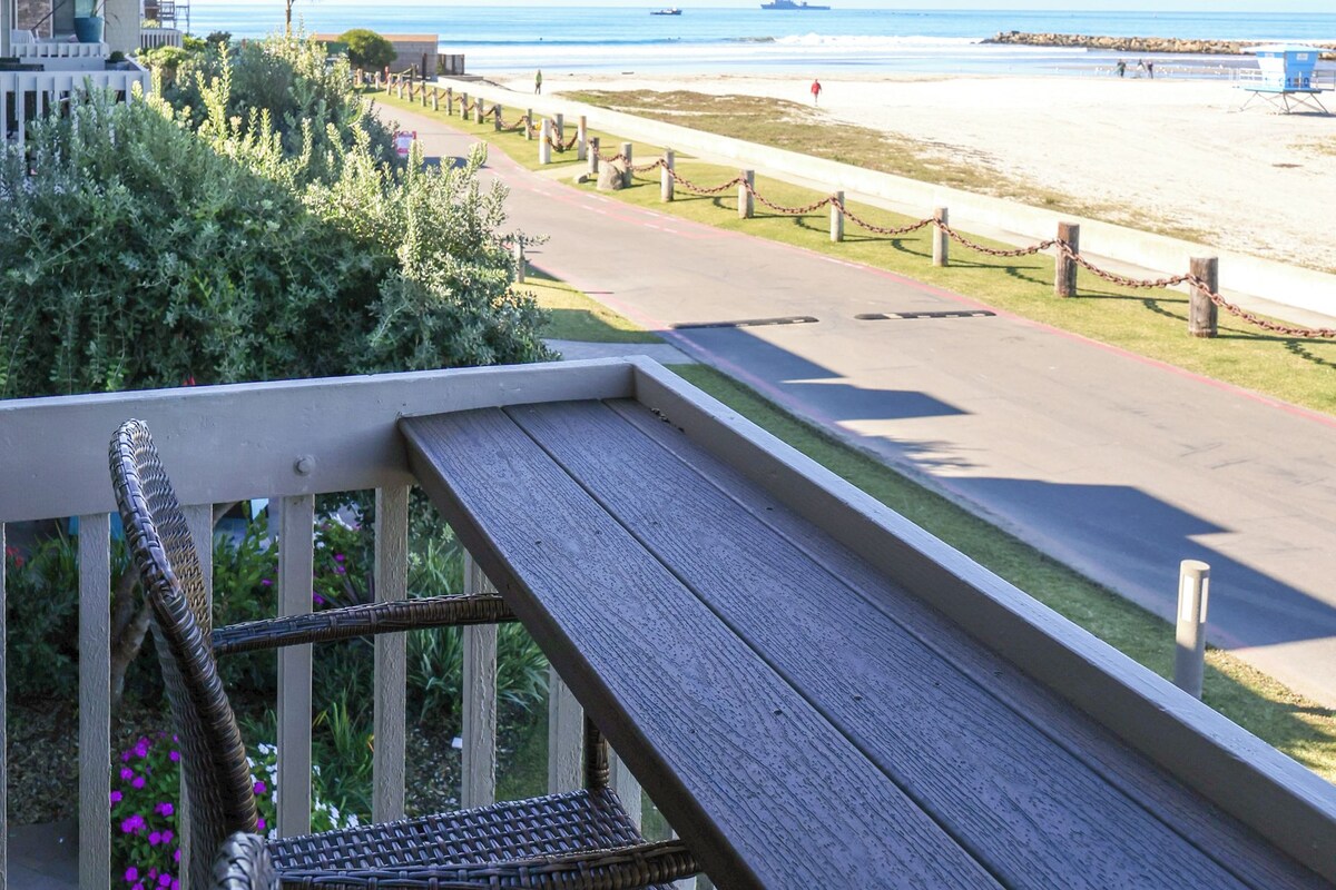 A balcony with a view of a beach, a paved walkway, and a wooden deck.