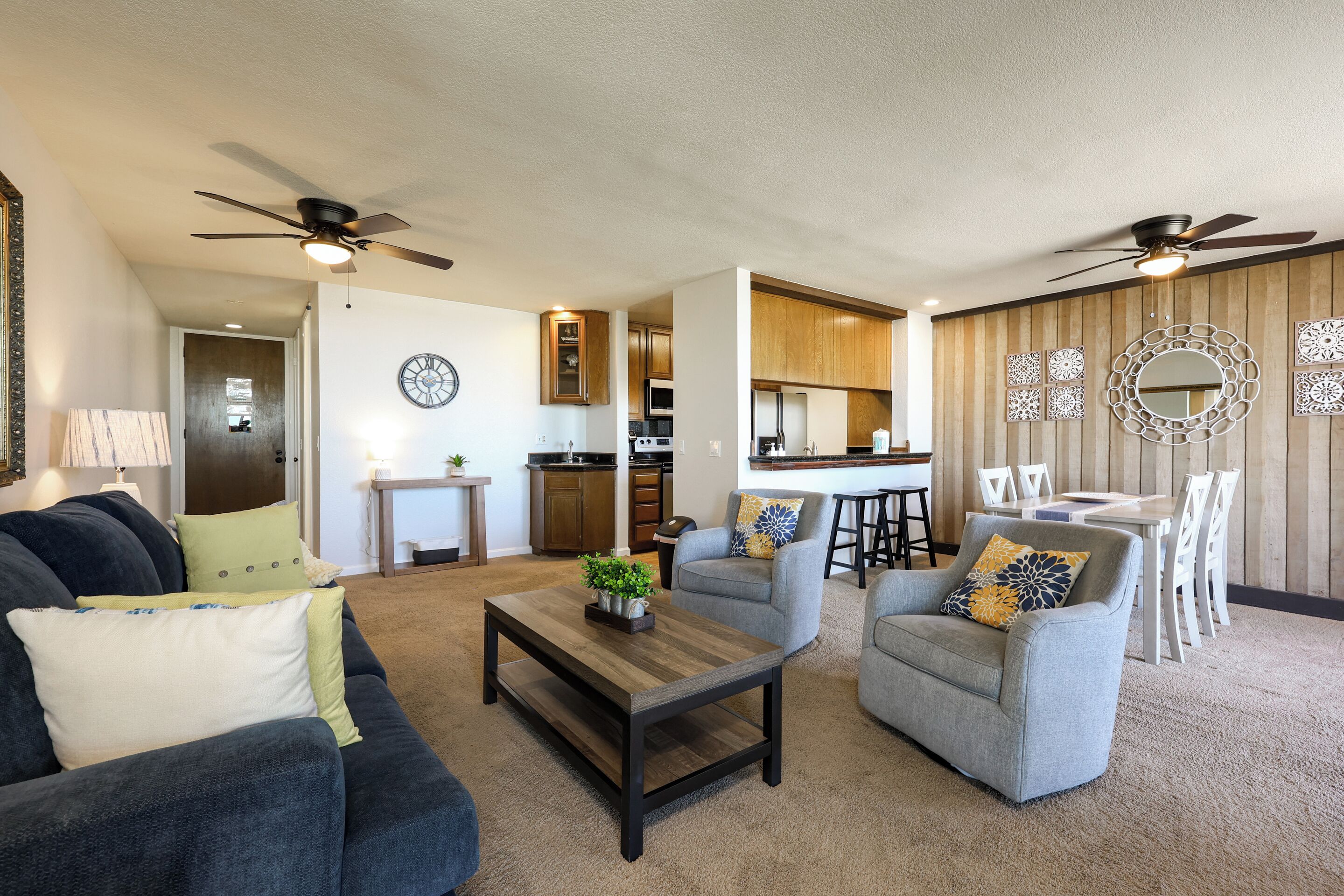 A living room with a dark blue sofa, a wooden coffee table, and two grey armchairs with decorative pillows. There is a kitchen in the background with wooden cabinets and a dining area with white chairs.