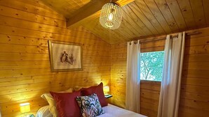 Un dormitorio de cabaña de madera con una cama, almohadas, una mesita de noche, una lámpara, un cuadro enmarcado en la pared y una ventana con cortinas.