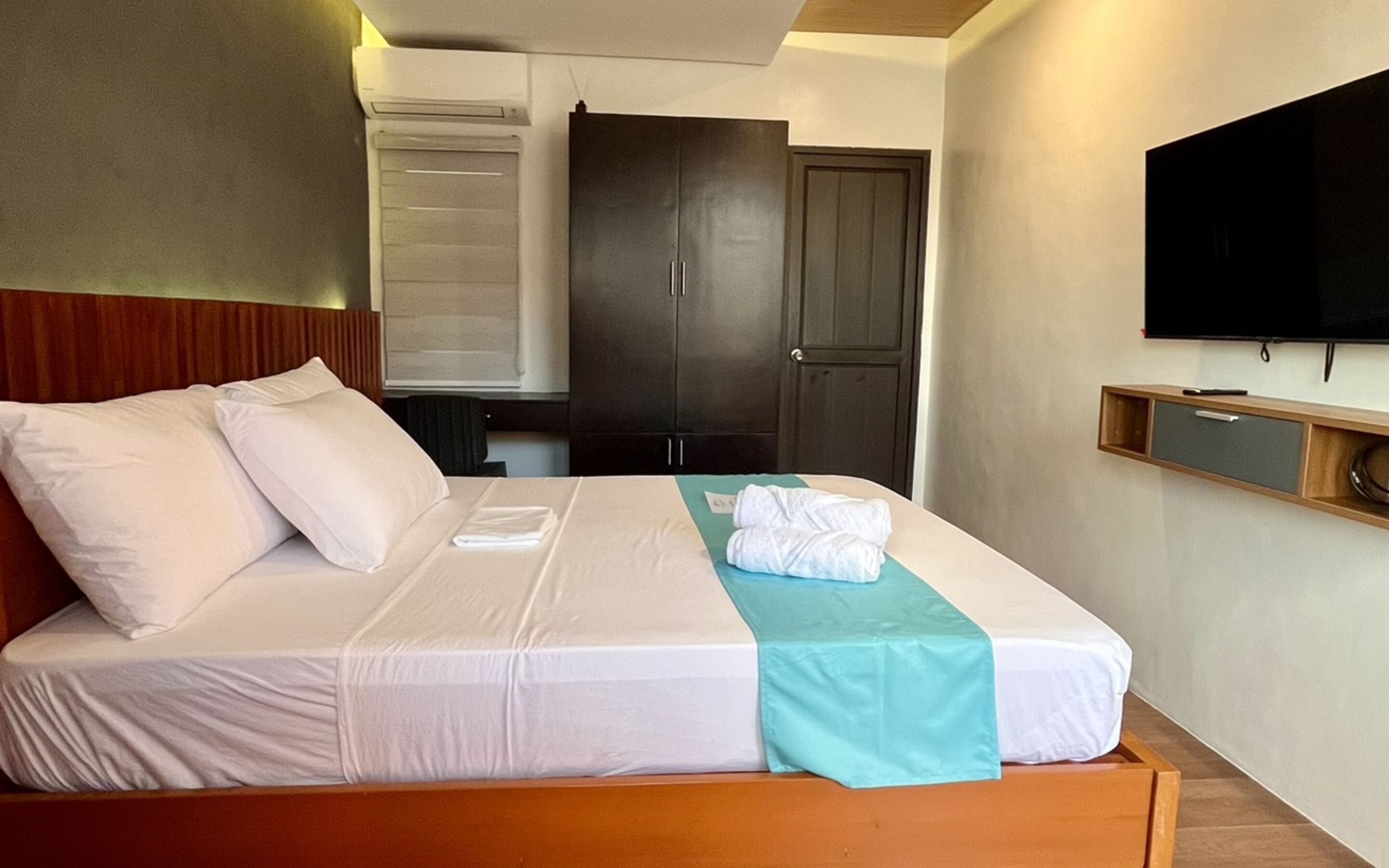A hotel room with a bed, a wooden headboard, a dark wardrobe, a flat-screen TV mounted on the wall, and a small wooden shelf with a pen and a bowl.
