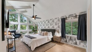 A bedroom with a large bed, a ceiling fan, a nightstand, a chair, and a window with curtains.