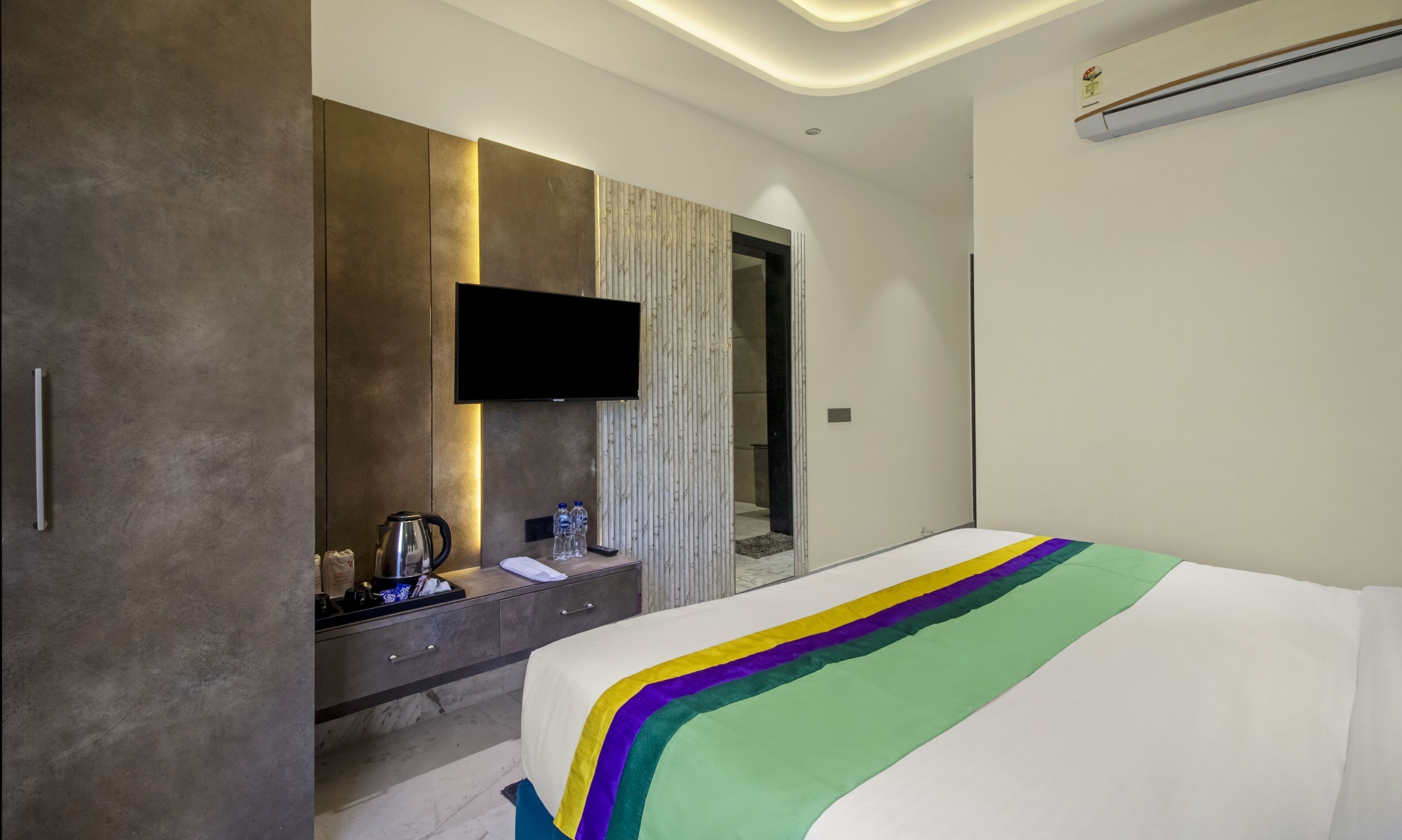 A modern hotel room with a large bed, a flat-screen TV mounted on the wall, a minibar, and a large mirror.