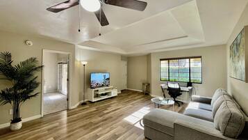 A living room with a ceiling fan, a TV on a white entertainment unit, a gray sofa, a coffee table, and a potted plant.