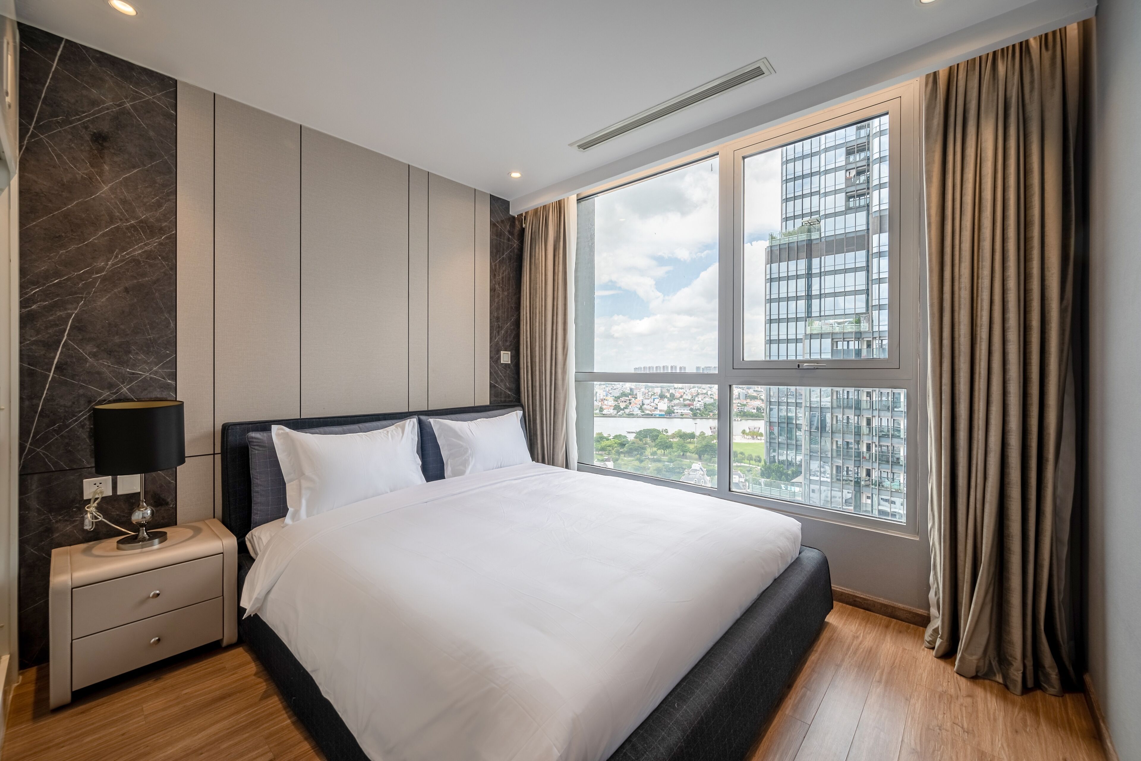 Une chambre d’hôtel moderne avec un grand lit, une table de chevet, une lampe et une vue sur la ville grâce à une grande fenêtre.
