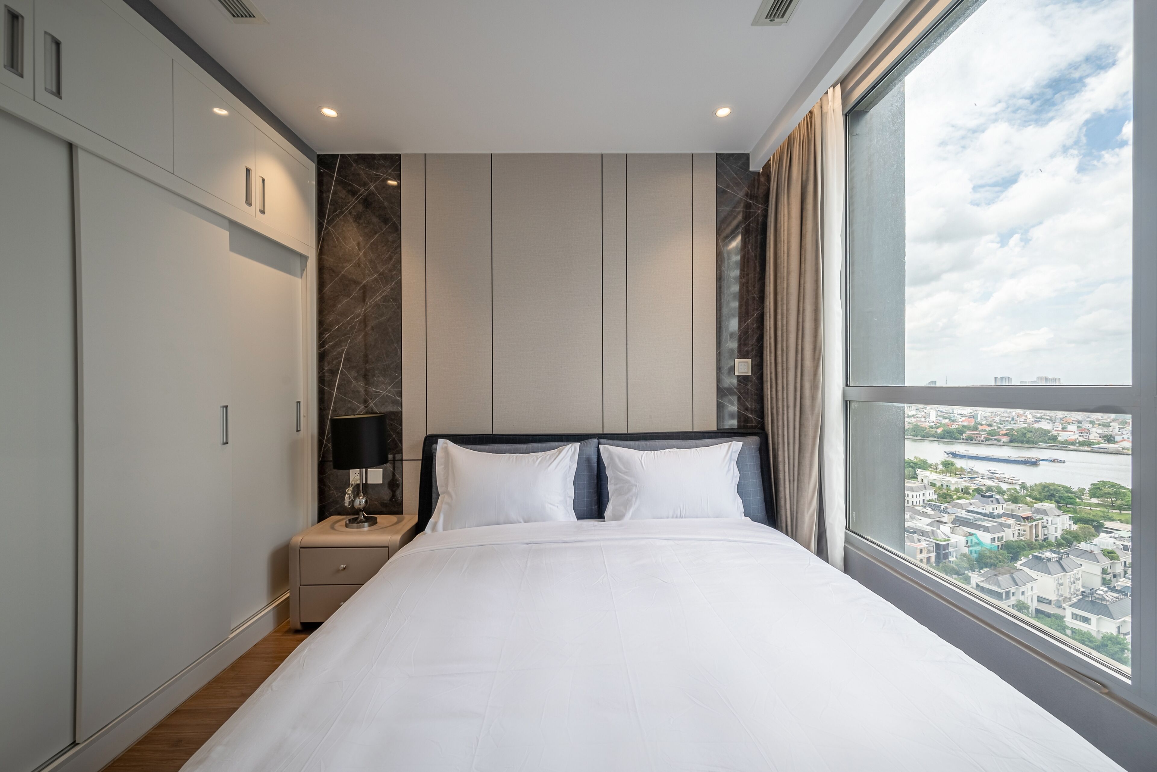 A modern bedroom with a large bed, a nightstand, a lamp, and a view of a cityscape through a large window.