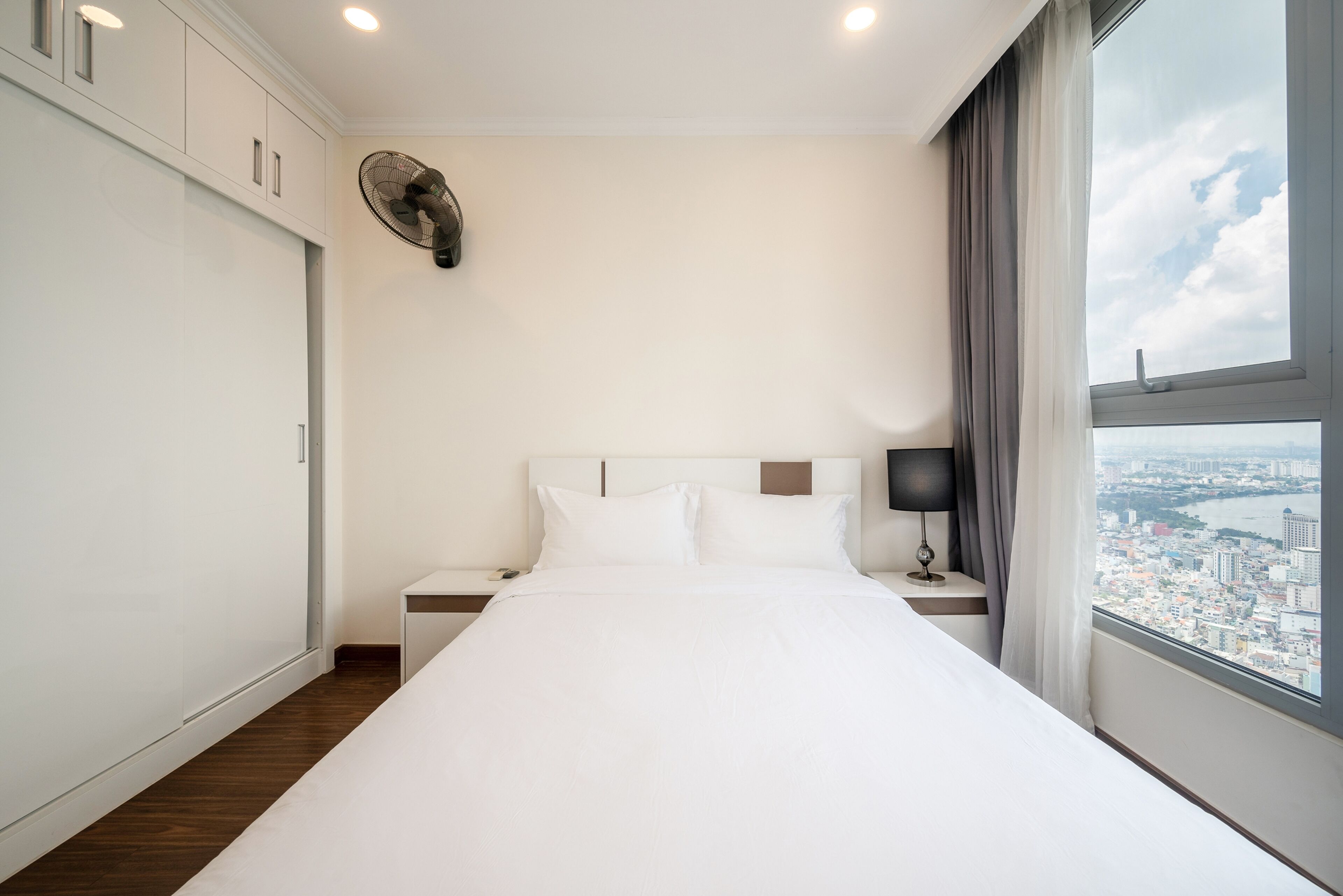 A modern hotel room with a large bed, a nightstand, a wall-mounted fan, and a window with a city view.