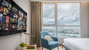 A modern hotel room with a large window offering a city view, a blue armchair with a colorful cushion, a flat-screen TV mounted on the wall, and a bedside table with books and a small plant. - The Celavie Serviced Apartment Landmark (Ho Chi Minh City)