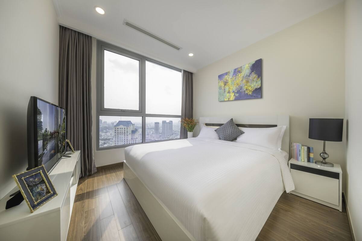 Ein modernes Hotelzimmer mit einem großen Bett, einem Fernseher, einem Nachttisch mit Büchern und einem Fenster mit Blick auf die Stadt.