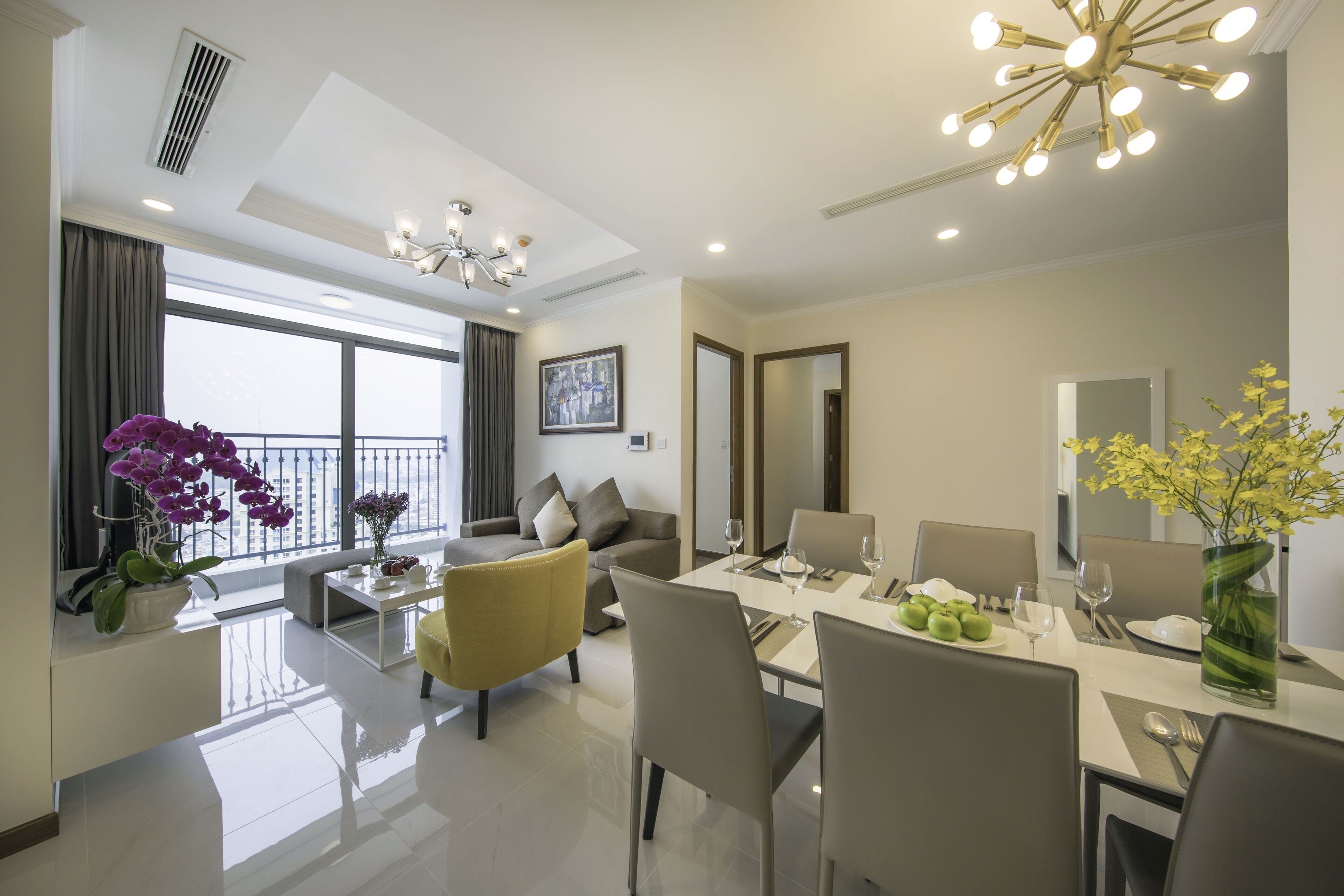 A modern hotel room with a dining area, a sofa, a yellow armchair, a large window with a view, and a chandelier.