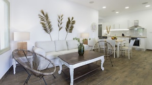 Una sala de estar moderna con un sofá blanco, dos sillones de mimbre, una mesa de centro de madera y una cocina al fondo.