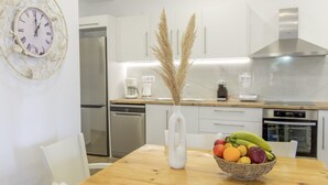 Una cocina moderna con armarios blancos, un refrigerador de acero inoxidable y una mesa de comedor de madera con un cuenco de frutas y un florero.