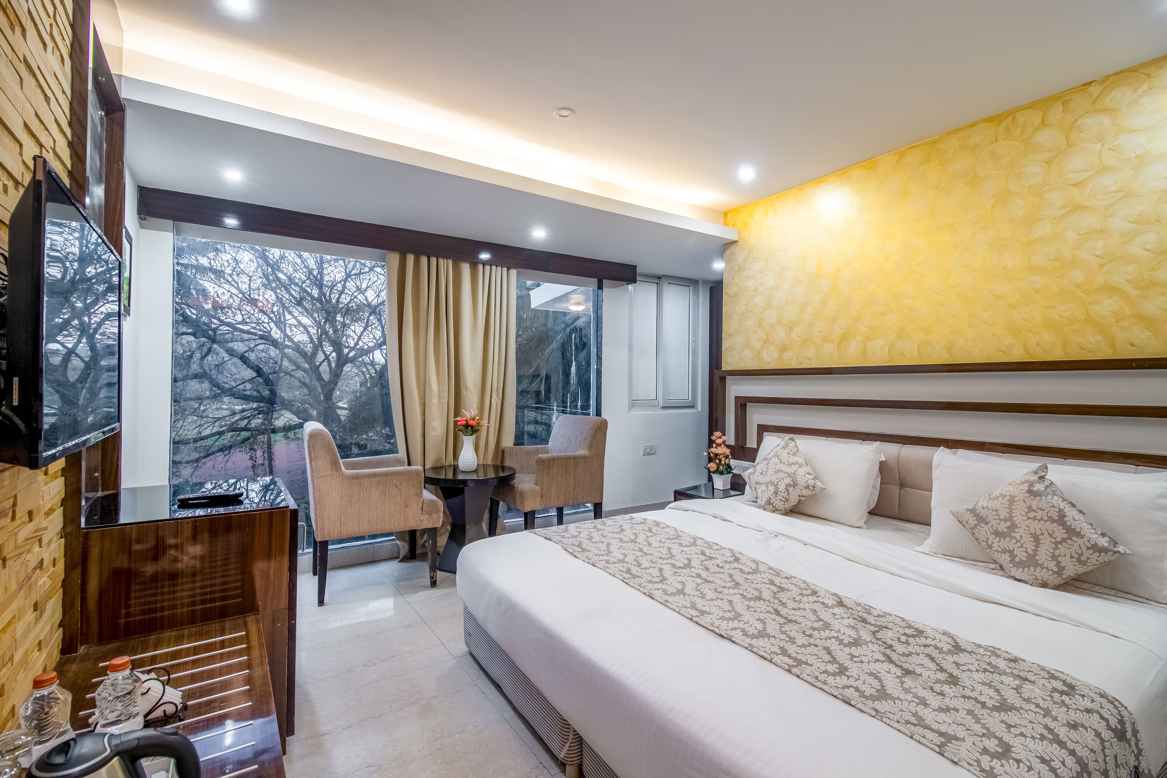 A modern hotel room with a large bed, a desk with chairs, a TV, and a view of trees through the window.