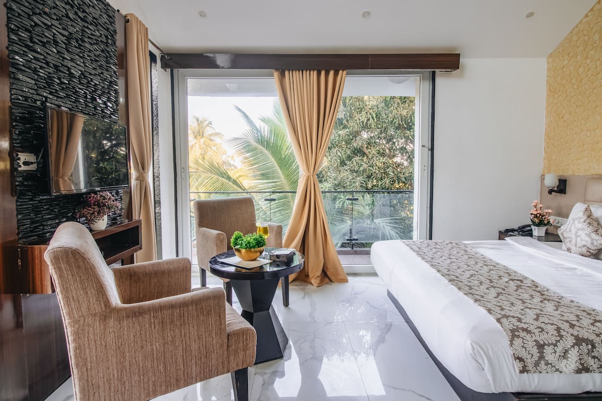 Quarto de hotel moderno com uma cama grande, área de estar com duas cadeiras, uma mesinha redonda com uma planta e uma varanda com cortinas.