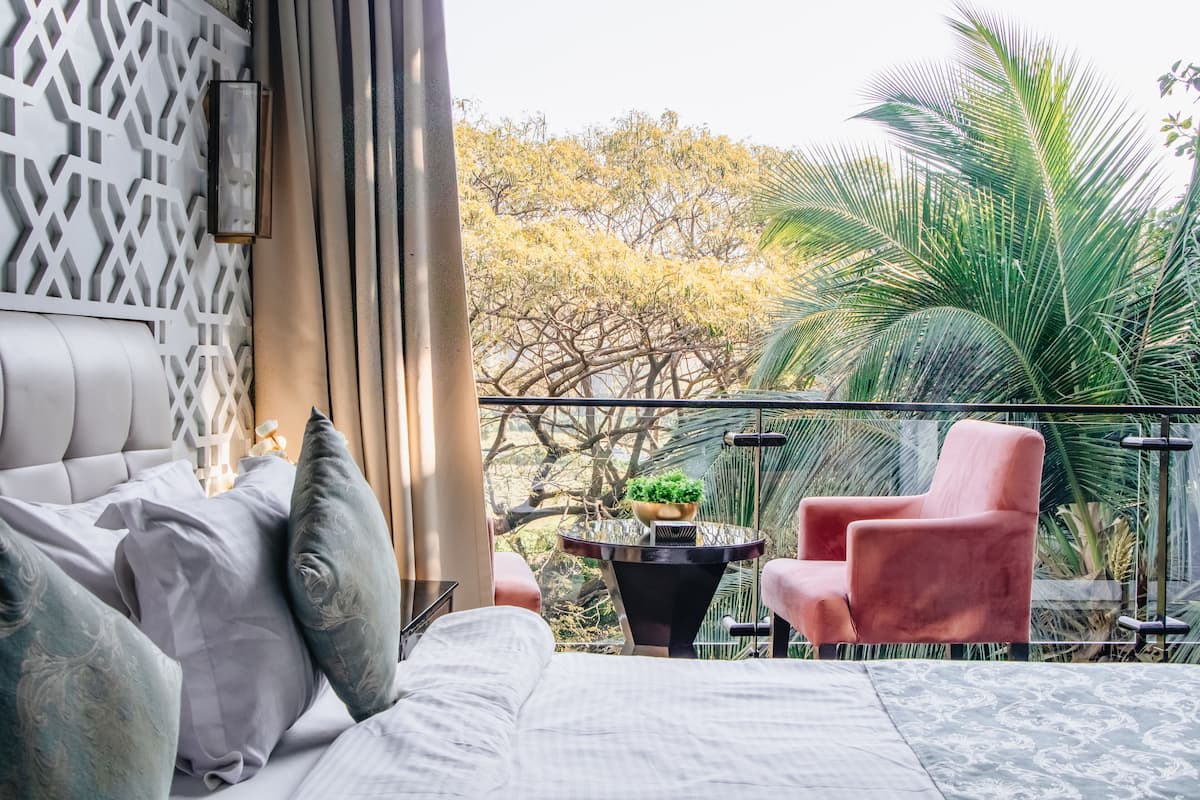 Quarto com cama, travesseiros, mesa de cabeceira, mesa com planta e uma cadeira. Há uma varanda com vista para árvores e uma planta de palmeira.