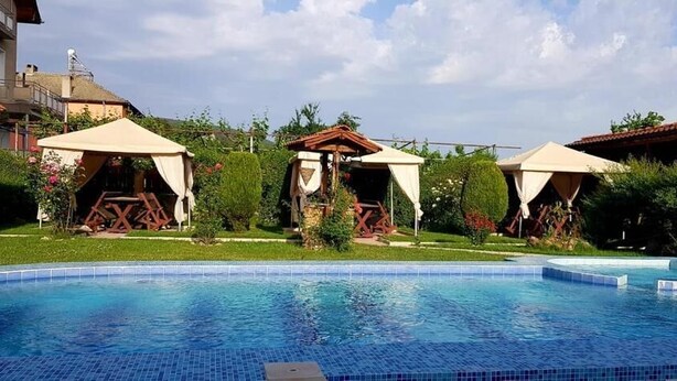 A swimming pool with a tiled edge, surrounded by greenery and a building with a red roof. - Sharkov Family Hotel  (Garmen)