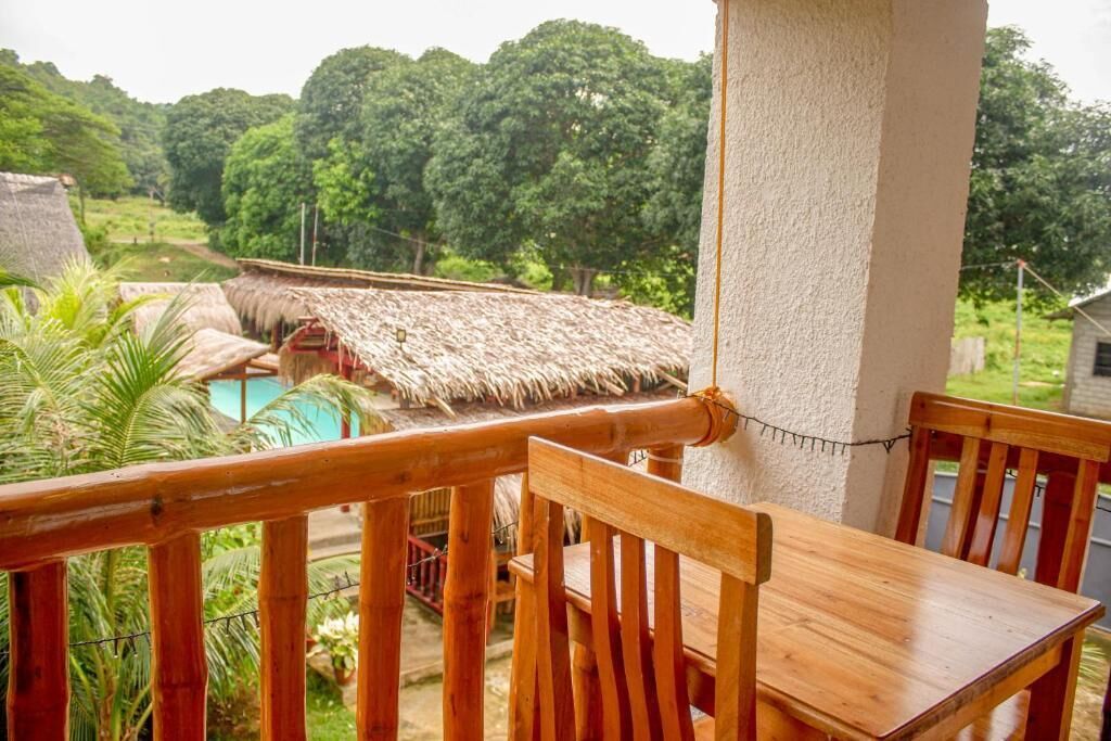 A wooden balcony with a thatched-roof structure in the background, a pool, and lush greenery.