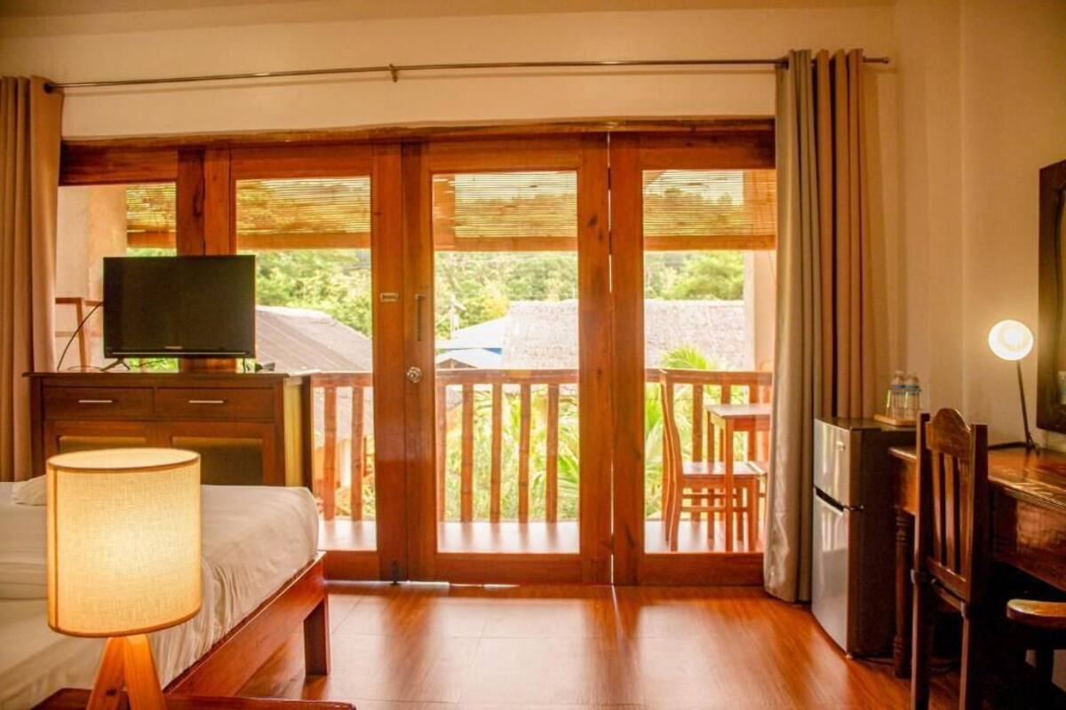 A hotel room with a bed, a desk with a computer, a television, and a balcony with a wooden railing.