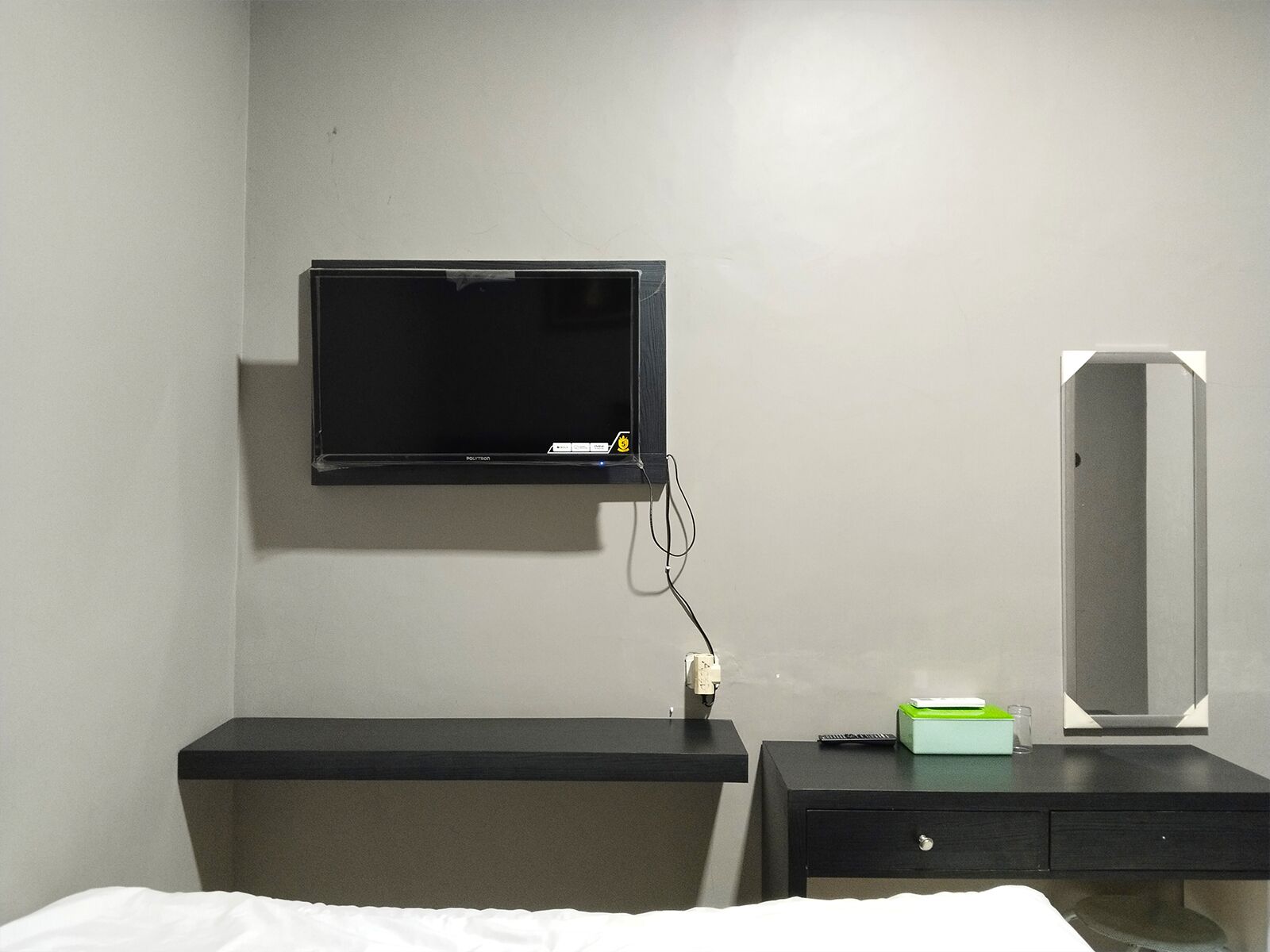 A hotel room with a bed, a desk with a mirror, a television mounted on the wall, and a small green box on the desk.