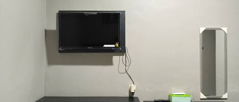 A hotel room with a bed, a desk with a mirror, a television mounted on the wall, and a small green box on the desk.