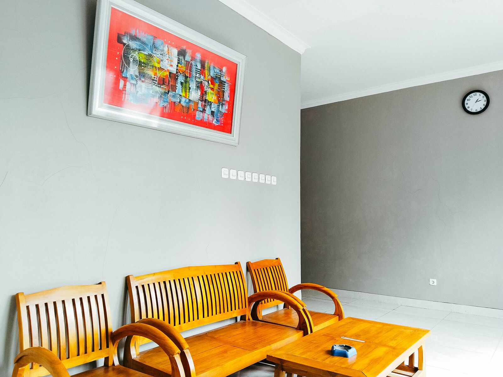 Una habitación con un banco de madera, una mesita con un control remoto, un reloj en la pared y un cuadro abstracto de colores.