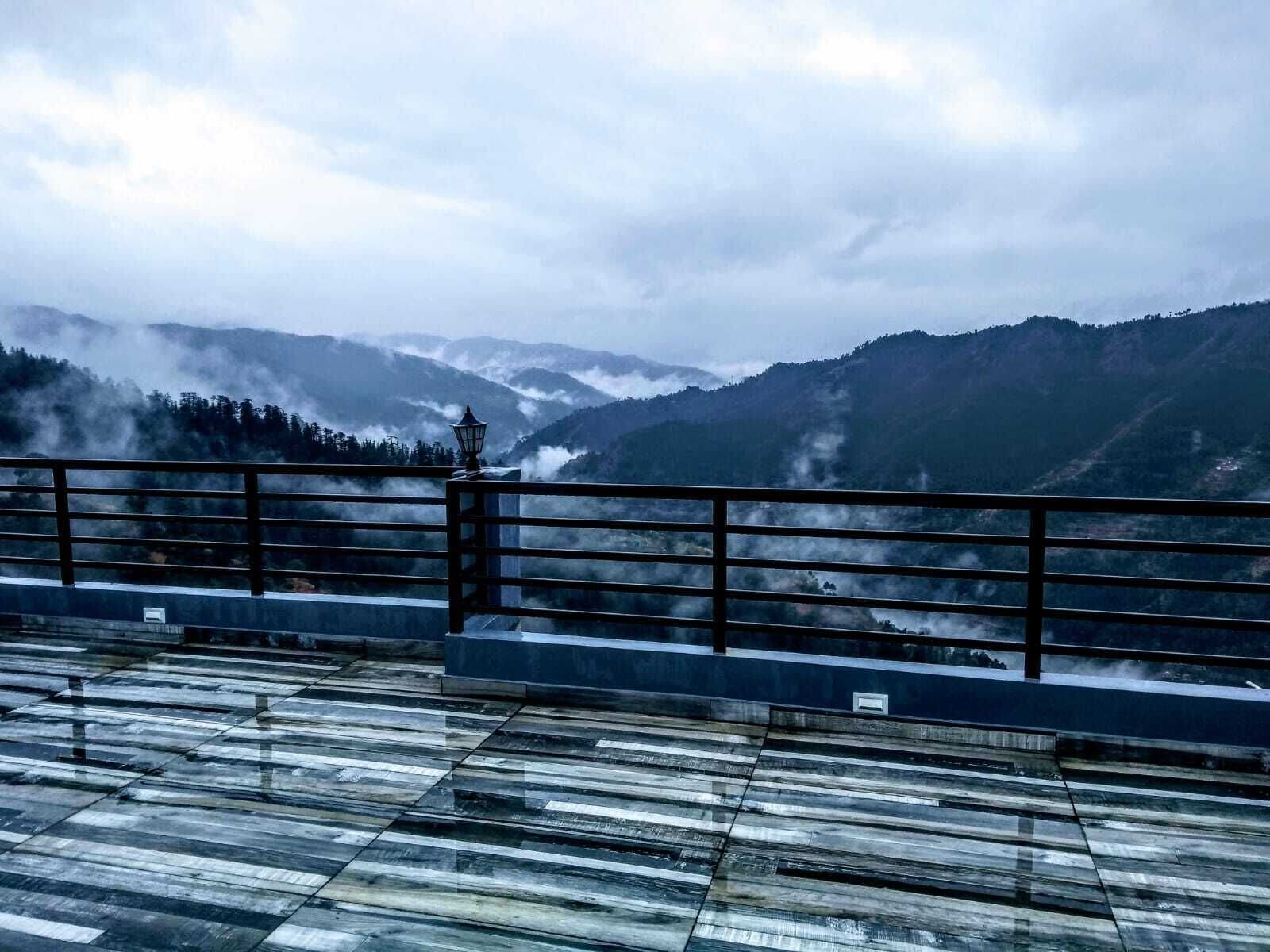 A view from a balcony with a metal railing, overlooking mist-covered mountains and a forested area.
