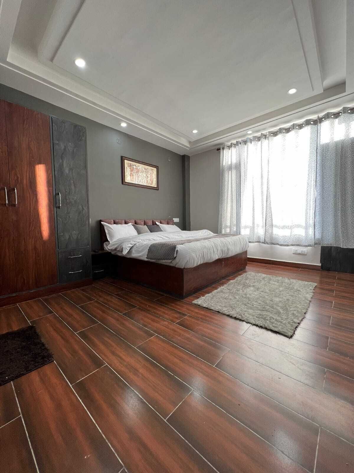 Ein modernes Schlafzimmer mit einem großen Bett, dunklem Holzboden, einem grauen Kleiderschrank und einem Fenster mit transparenten Vorhängen.