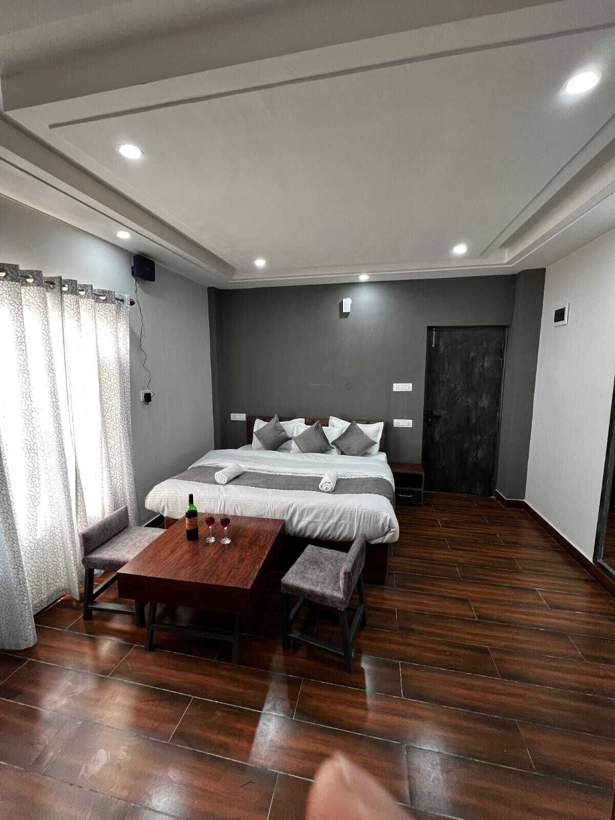 A modern hotel room with a large bed, a small table with a bottle and glasses, and a wooden floor.