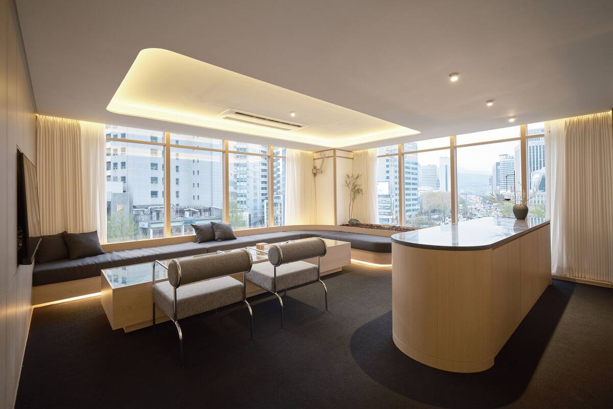 Een moderne woonkamer met een bank, een salontafel en een gebogen houten tafel. Grote ramen bieden uitzicht op de stadsgezicht.