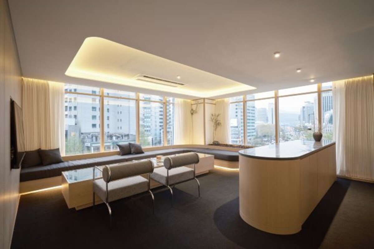 Een moderne hotelkamer met een gebogen receptiebalie, een zithoek met banken en een bijzettafeltje, grote ramen met uitzicht op de stad, en een minimalistisch design.