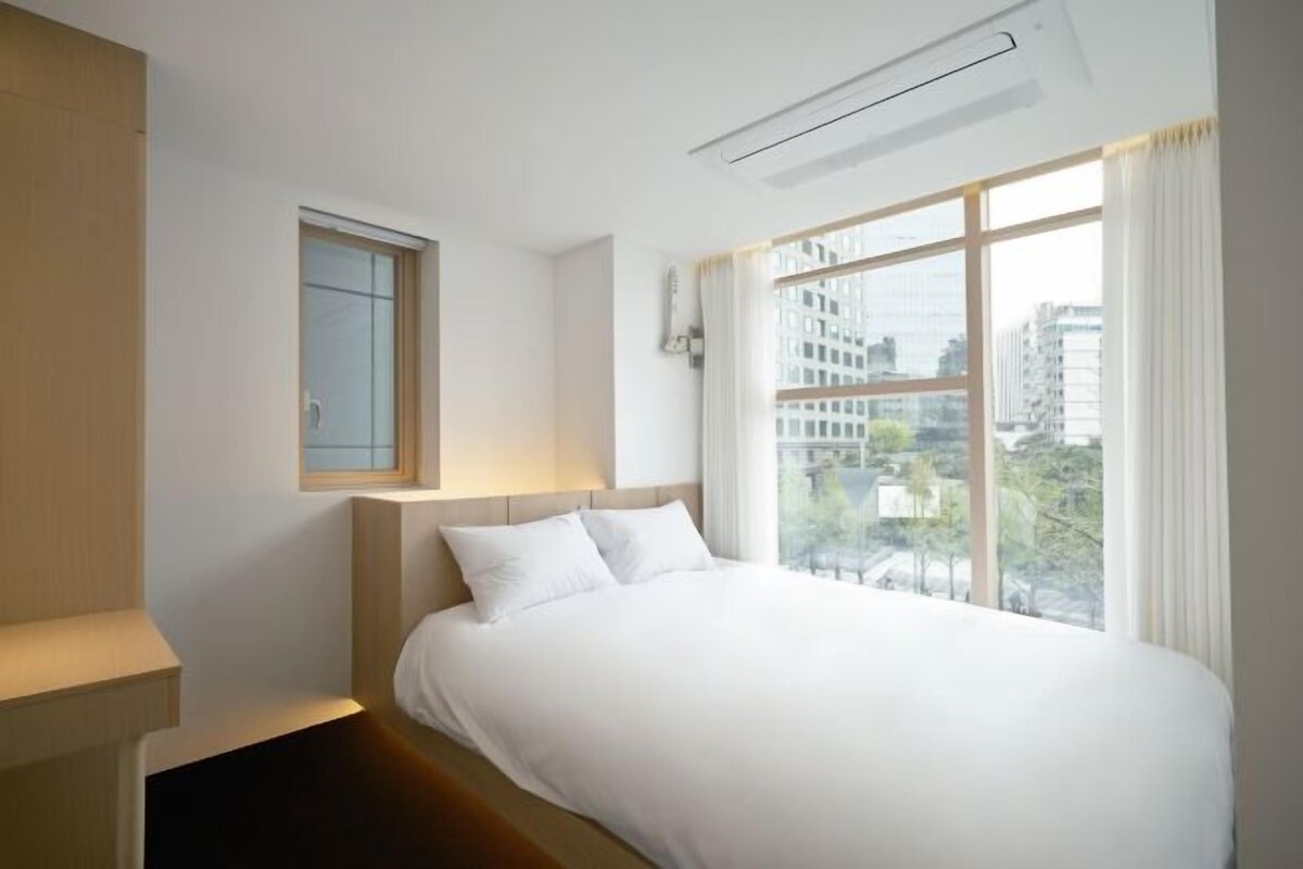 Ein Hotelzimmer mit einem großen Bett, einem Fenster mit Blick auf die Stadt und einem kleinen Nachttisch.