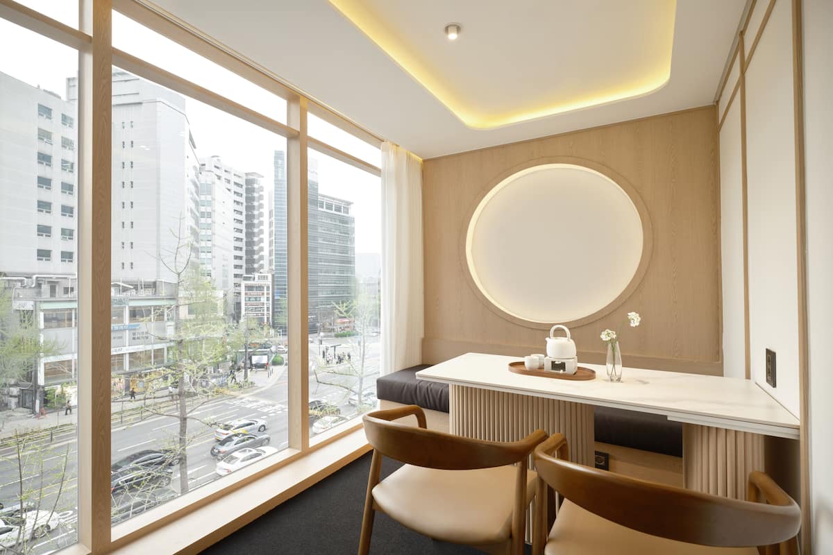 Een moderne hotelkamer met een groot raam, een ronde spiegel, een klein tafeltje met een waterkoker en twee stoelen.