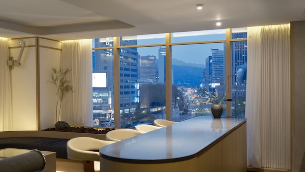Une chambre d’hôtel moderne avec un comptoir d’accueil incurvé, un coin salon avec des fauteuils, une grande fenêtre donnant sur la ville et une petite plante posée sur le rebord de la fenêtre.