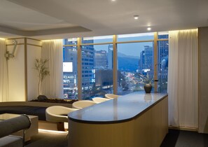 Ein modernes Hotelzimmer mit einer gebogenen Rezeption, einer Sitzecke mit Stühlen, einem großen Fenster mit Blick auf die Stadt und einer kleinen Pflanze auf der Fensterbank.