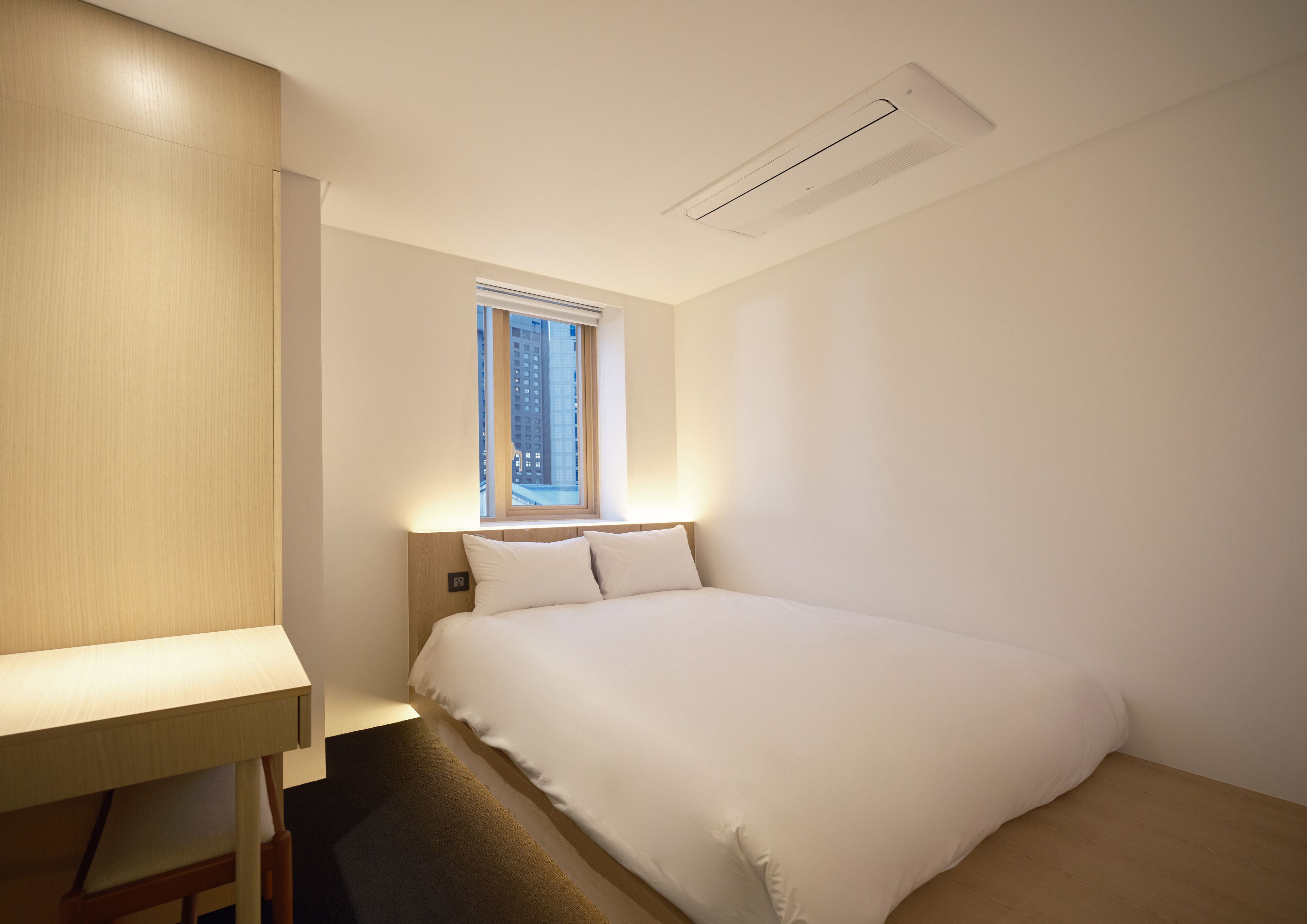 A modern hotel room with a neatly made bed, a desk, and a window with a view of tall buildings.