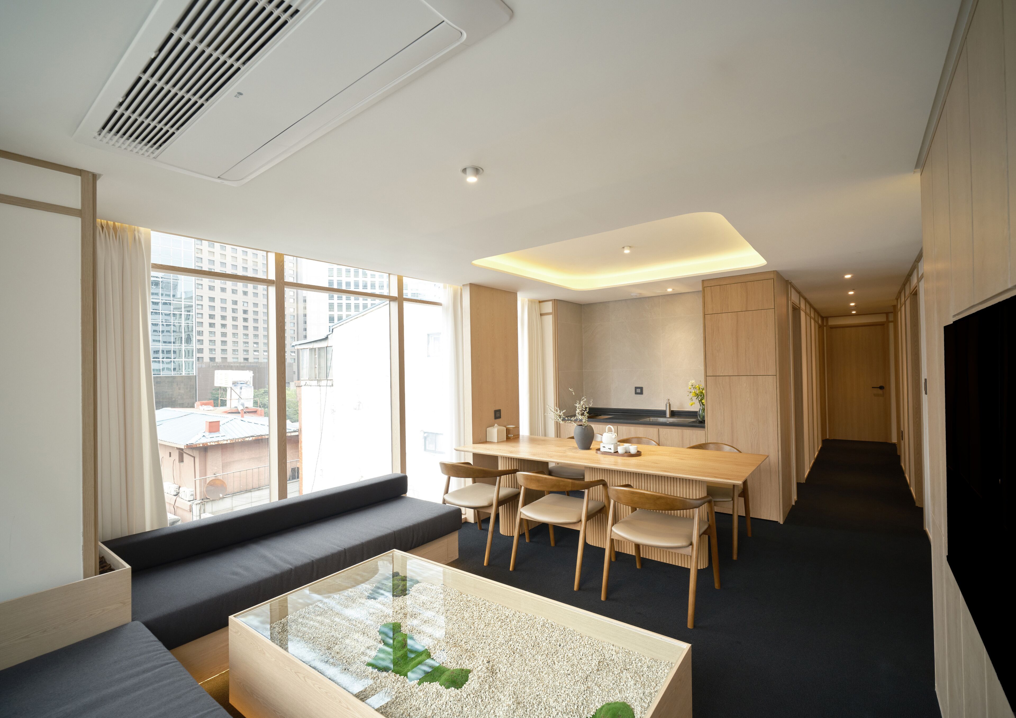 A modern hotel room with a large window, a sofa, a dining area with chairs, and a kitchen area.