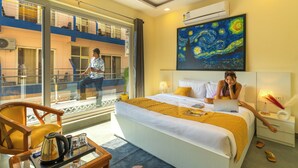 A hotel room with a bed, a desk, a chair, a window, and a painting on the wall. - The Social Stays Rishikesh (Narendranagar)
